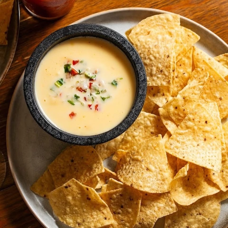 Crispy tortilla chips surround a bowl of creamy queso topped with diced peppers, alongside a red drink on a wooden table.
