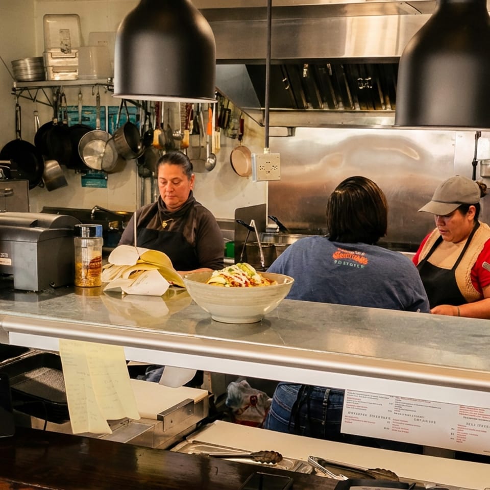 Three women working behind a busy restaurant counter with food and kitchen equipment.