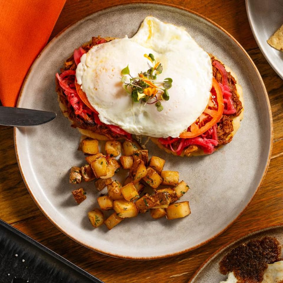 Breakfast plate with two fried eggs, shredded meat, pickled onions, tomato, and crispy home fries.