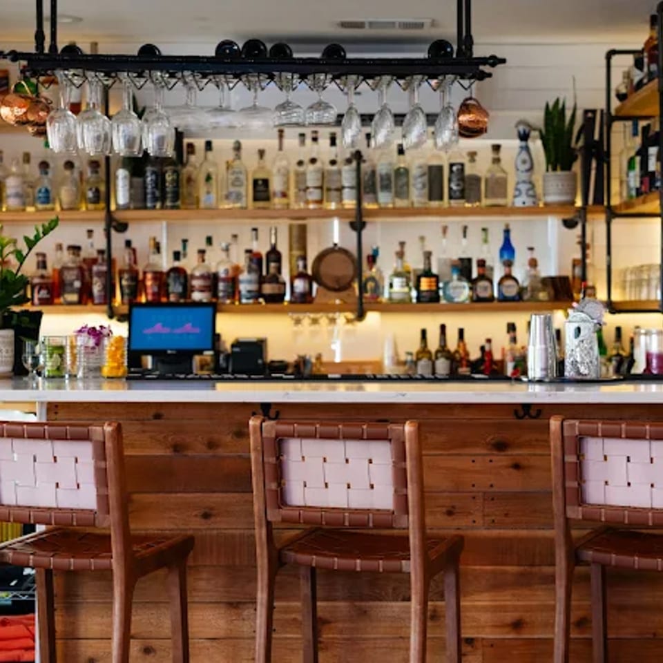 Warm, inviting restaurant bar with wooden stools, backlit liquor shelves, hanging glassware, plants, and a computer terminal.