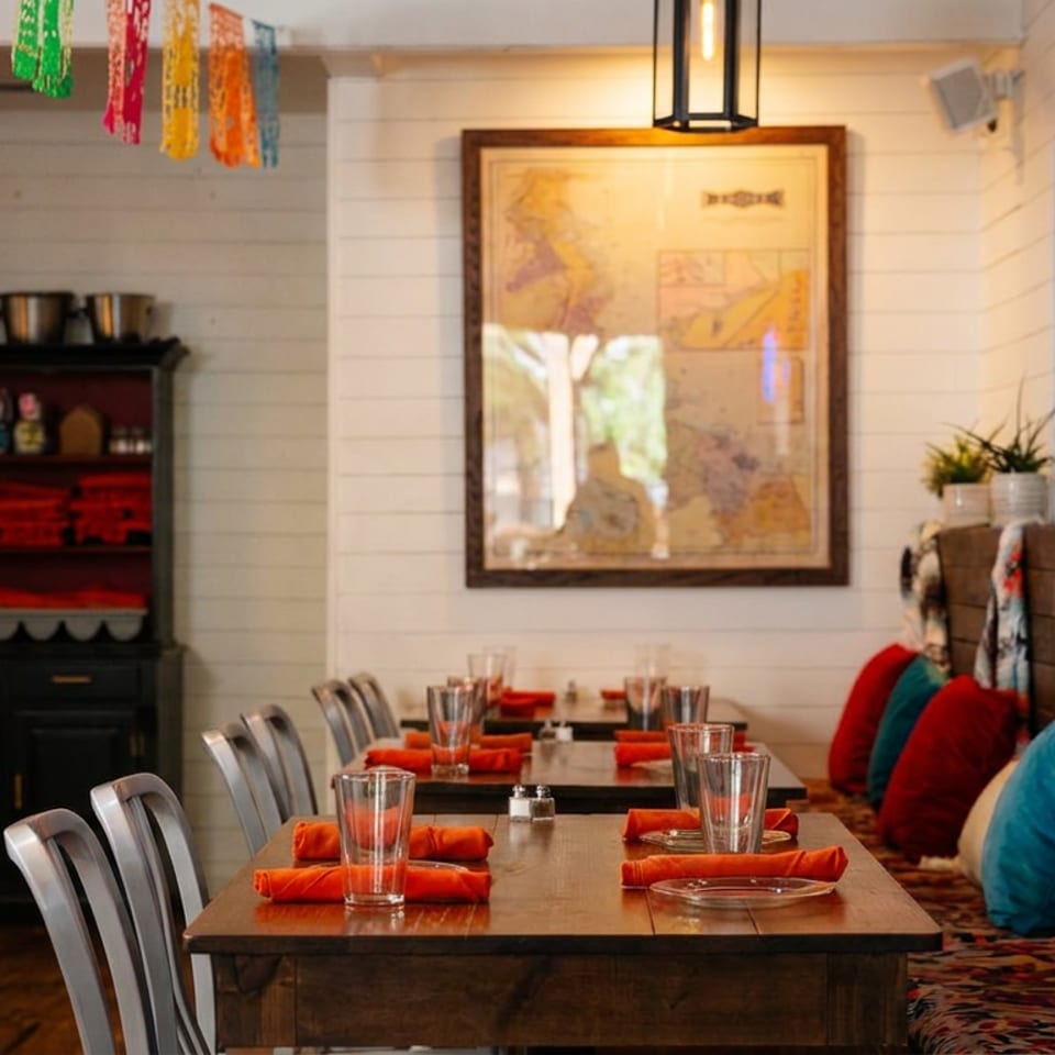 Restaurant interior with wooden tables, metal chairs, colorful cushions, and a large framed map on the wall.