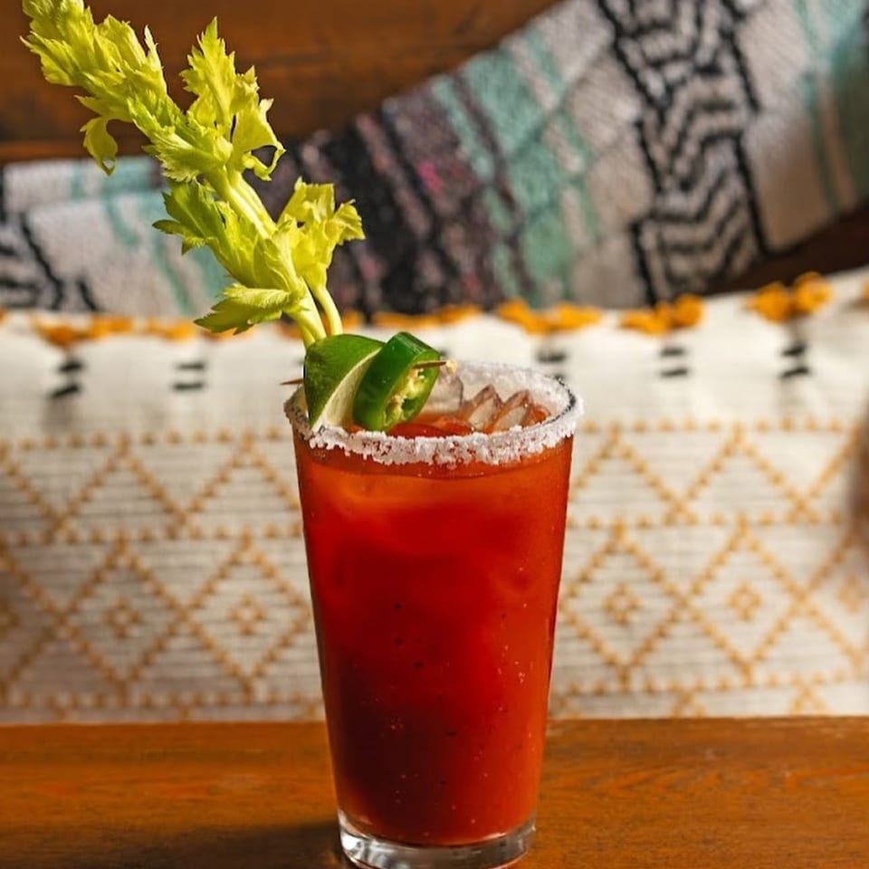 Red Bloody Mary cocktail with a salt-rimmed glass, lime wedge, jalapeño, celery, and savory garnishes on a restaurant table.