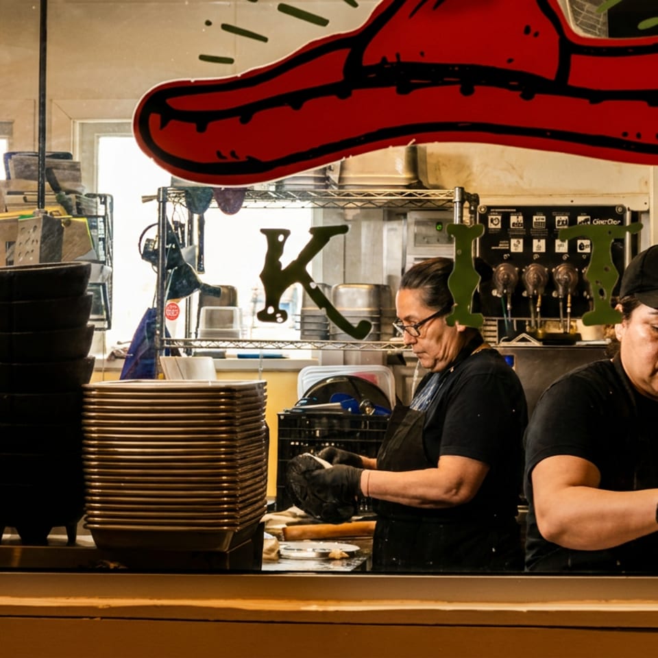 Two women chefs in black uniforms working in a busy restaurant kitchen viewed through a window.