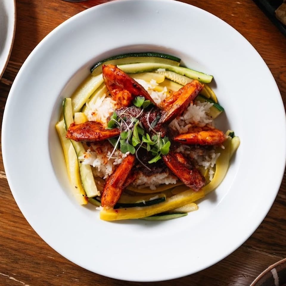 White plate of rice topped with grilled shrimp, microgreens, and colorful zucchini strips on a wooden table.