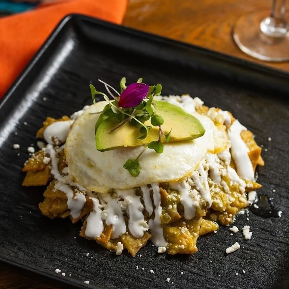Layered chilaquiles topped with a fried egg, avocado, crema, crumbled cheese, and microgreens on a black plate.