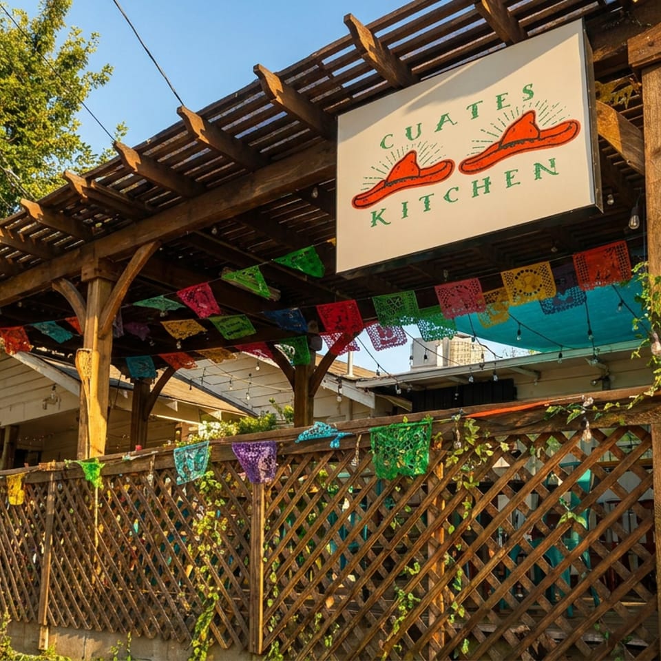 Exterior of Cuates Kitchen, a Mexican restaurant with a wooden pergola, colorful papel picado, and a fence.