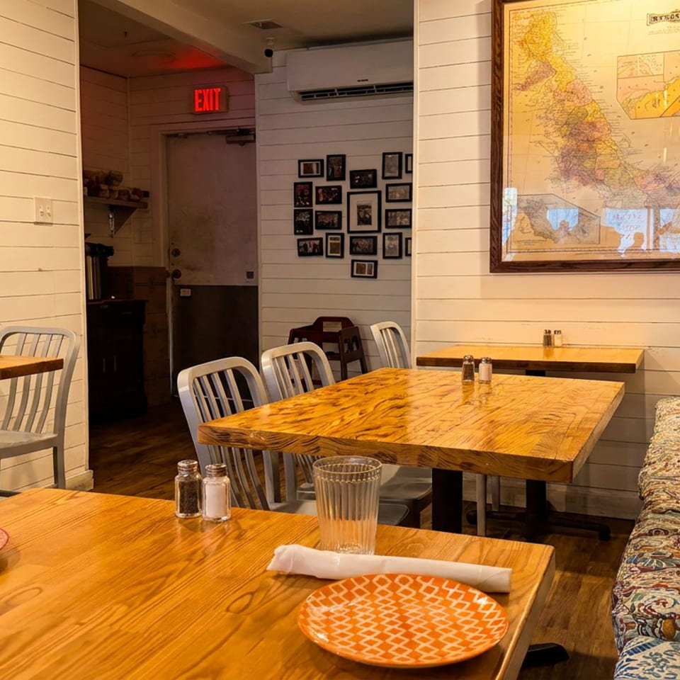 Cozy restaurant interior with wooden tables, white shiplap walls, a map, and a cushioned bench with pillows.