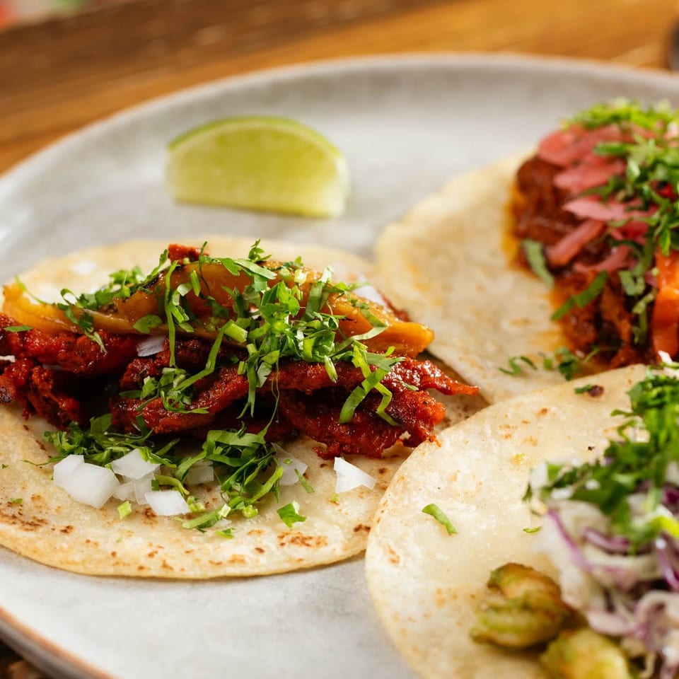 Three soft tacos filled with spiced meat, pickled vegetables, cilantro, onions, and lime on a plate.