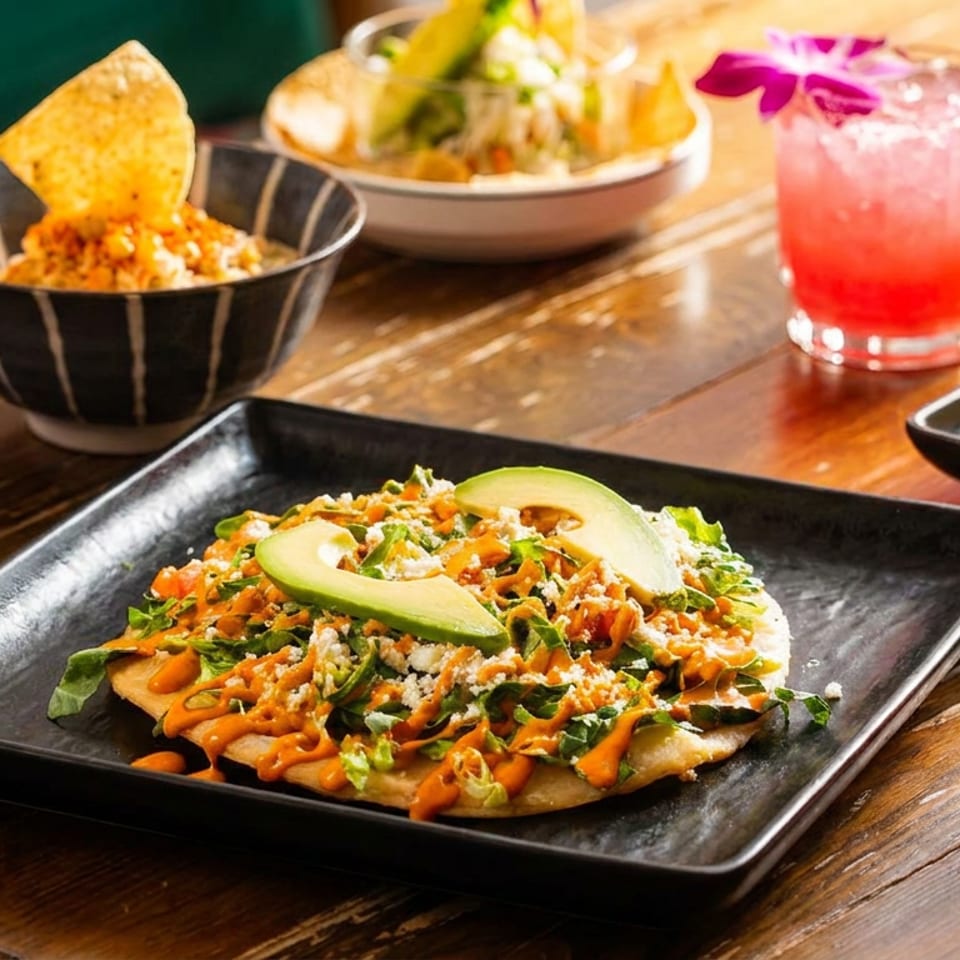 A vibrant spread of Mexican dishes, including a tostada, guacamole, and colorful cocktails on a wooden table.